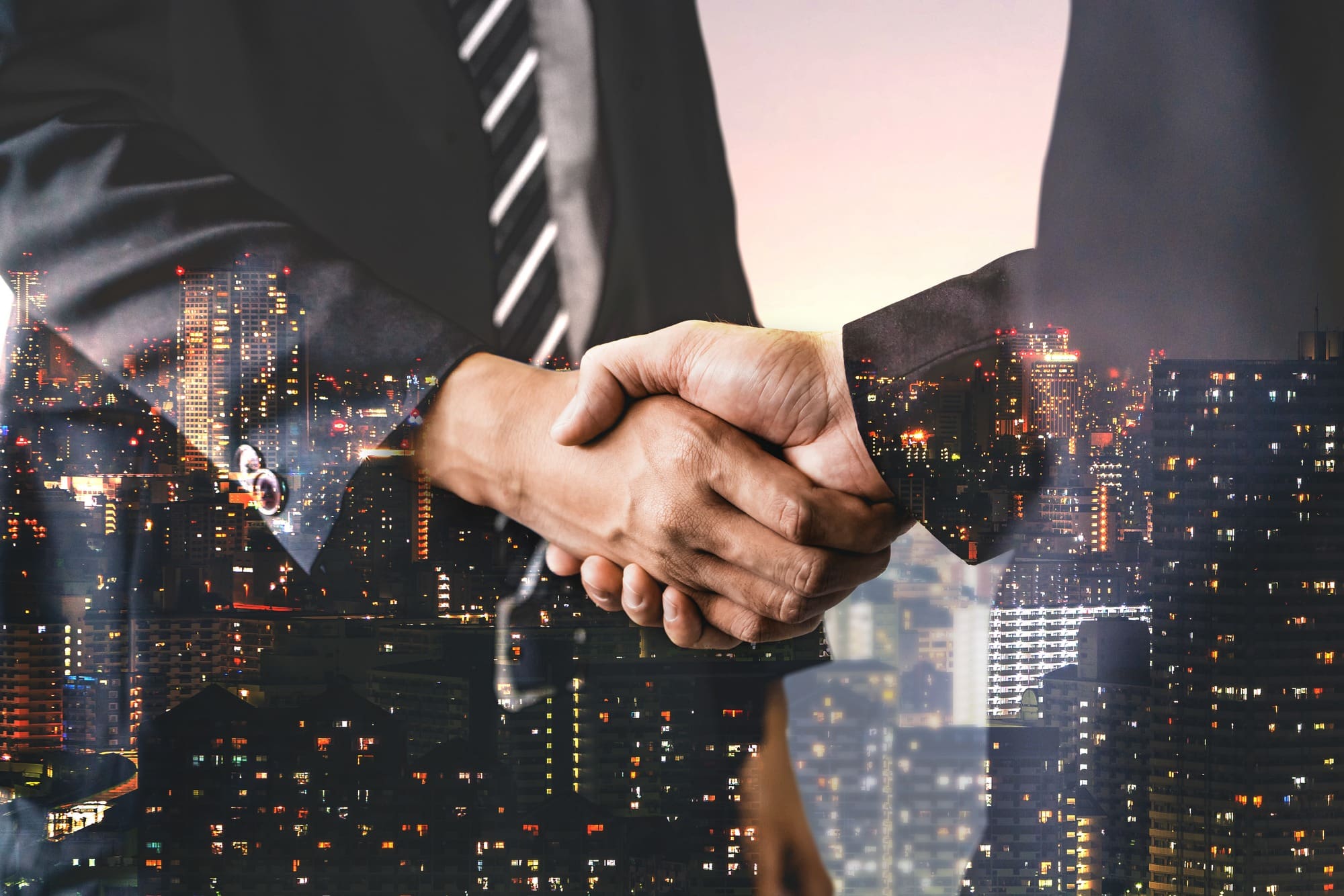 Double exposure image of business people handshake on city office building in background showing partnership success of business deal.
